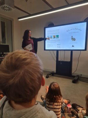 Frau zeigt einer Gruppe von Kindern das Buch "Nelly und die Berlinchen" auf einem Whiteboard